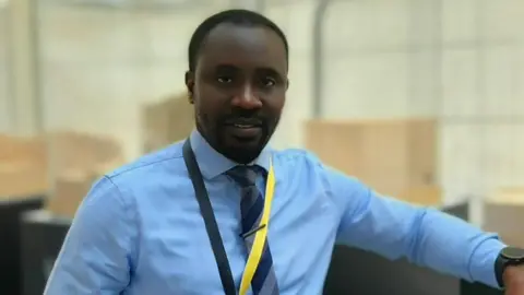 Titi Owode Titi looking at the camera, wearing a smart blue shirt, a striped tie and trousers.