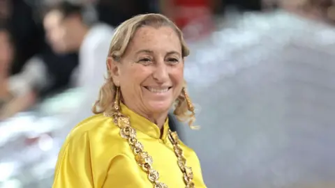 Getty Images A smiling Miuccia Prada wears a yellow satin top and large gold chain at the Met gala