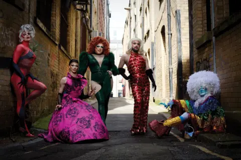 Magnus Hastings Five drag queens pose in an alley way. One in a red jumpsuit covered in handprints, one in a pink floral gown, another in a green sparkly jumpsuit, a fourth in a red dress decorated in fish scales and the fifth in a rainbow tasseled outfit with a white feathered afro wig.