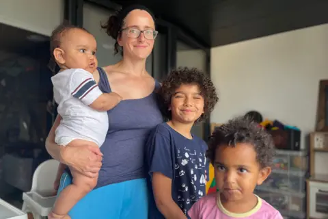 BBC A woman with curly brown hair and glasses, Thea, with her three children: Isaac, a baby, JJ, aged two, and Moses, 9