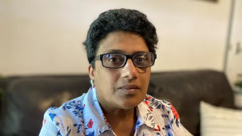 BBC Kumudini Nicola sitting on a sofa in her home. She is wearing glasses and tears are in her eyes.