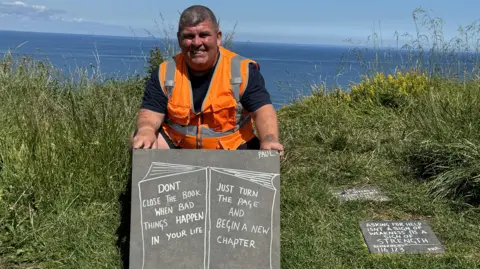 BBC A man wearing an orange Hi-Viz bib and holding a slate sign next to a cliff edge