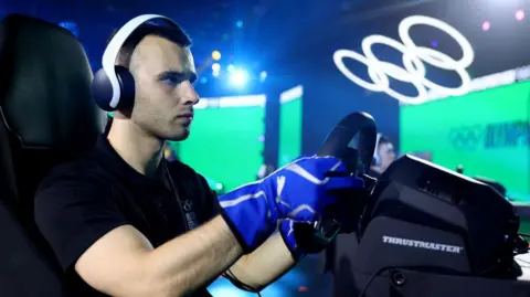 Getty Images  A young man wearing white over-ear headphones sits in front of a videogame controller steering wheel in an arena setting. He wears an expression of concentration as two large screens glow green behind him and a large set of white Olympic rings lit in white hovers over the stage