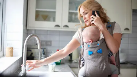 Getty Images A woman pours a glass of water while carrying her baby in a sling