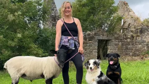 SWNS Robyn with a sheep and two dogs in front of a ruined building