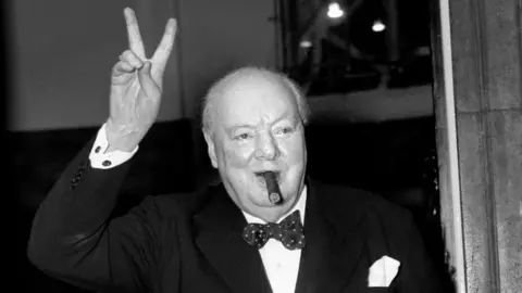 PA Media Winston Churchill, in a suit and bow tie and with a cigar in his mouth, makes V for Victory sign with his right hand 