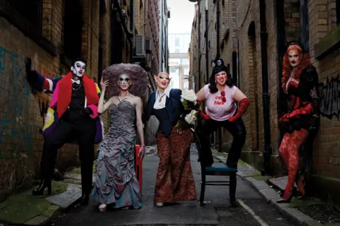 Magnus Hastings Five drag performers pose in an alley. The first, dressed as a man wears a white face and multi-coloured coat. The second has a grey dress with a large round hate. The third wears a blazer and wide trouers, the third stands on a chair in a t-shirt and red elbow-length gloves. The last rests with her back on the wall wearing red patterned tights and a corset.