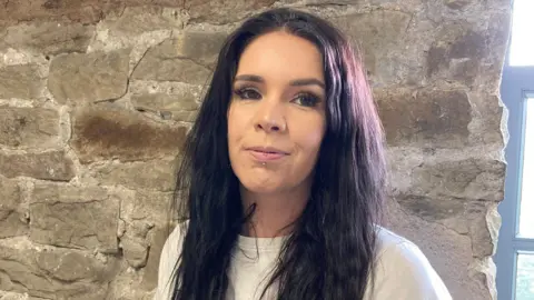 BBC Key Crawford wearing a white t shirt with piercing on a lip standing in front of a stone wall next to a window. Key has long dark hair.