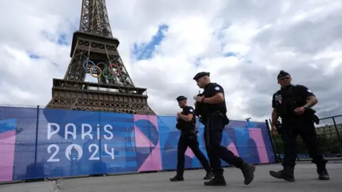 PA Media Police officers walk in front of the Eiffel Tower in Paris. The Olympic rings can be seen mounted half way up the tower.