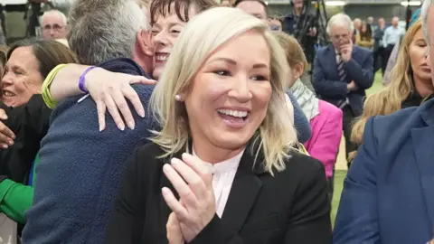 PA Sinn Féin deputy leader Michelle O'Neill clapping and smiling broadly. She is wearing a black jacket over a white shirt and has medium-length blonde hair