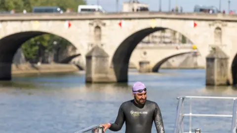 ANDRE PAIN/EPA-EFE/REX/Shutterstock A swimmer emerging from the Seine in Paris wearing a wetsuit and a lavender swimming cap, with goggles over the top of it