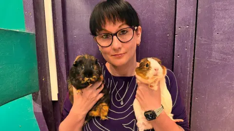 Masons' Cavies A woman with short black hair and glasses holds two guinea pigs while looking at the camera