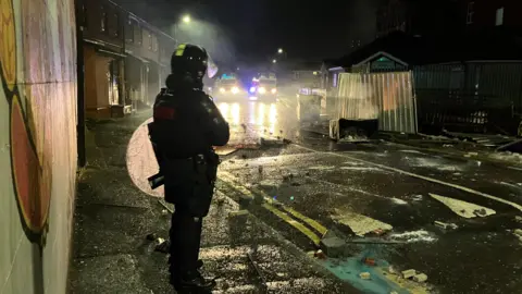 Single policeman in riot gear standing on a dark street surrounded by debris, with two police vans in the background with their lights on 