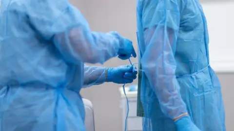 Getty Images A surgeon in an operating theatre, dressed in full scrubs, helps a colleague, dressed in the same manner, tie his scrubs