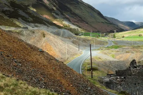 Jon Pountney Piles of different coloured rubble and rock at the side of the road going through a valley