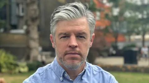 Stuart Antrobus/BBC A man with grey hair worn in a side-part looks at the camera. He is stood in a park and is wearing a blue shirt with the collar unbuttoned.