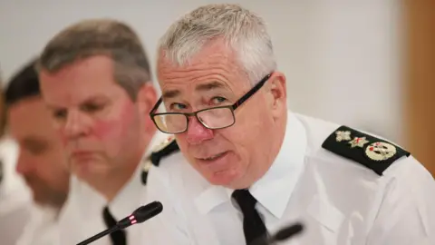 PA PSNI Chief Constable Jon Boutcher, in uniform, leans over a mic at a conference