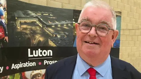 Amy Holmes/BBC John Tizard smiling in front of a sign that says "Luton"