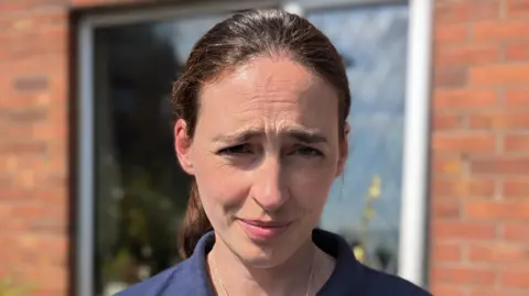 A woman with brown hair which is tied back in a ponytail looks at the camera while standing in front of a red brick house