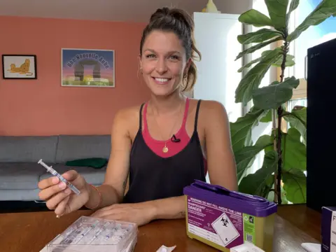 Tracey Langford / BBC Egg Donor Charlotte Holmes smiles while holding a hormone injection in her living room