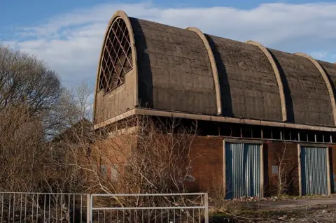 Jon Pountney A high arched roof building with with corrugated iron covering doorways, and rubble outside