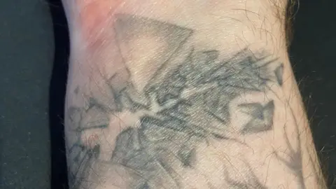 A close up image of a man's wrist with faded shards of glass tattooed around a faint horizontal scar