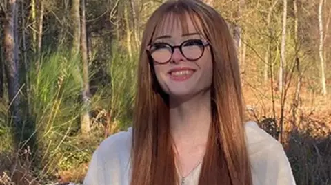 Family handout Brianna Ghey, with long auburn hair and round glasses, smiles and wears a white zip-up top in front of autumnal woodland and snowy ground