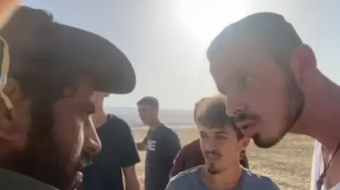 Jordan Valley Activists Ahmad, wearing a hat, appears to be harassed by settlers. It shows a close up side profile of Ahmed's face, with a man appearing to talk into his face. A number of other men are gathered around them.