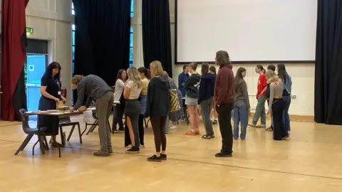 Pupils at Kingsbridge Community College collect their A-level results in three queues from staff stood behind tables in a sports hall