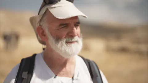 Gil Alexander standing in the sun, shown from the shoulders up. He has a cap on his head, and is looking off camera.