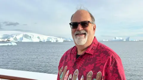 David Austin MD Man wearing bright red shirt with surf boards and flowers on it, wearing sunglasses. Standing in front of icebergs and the sea.