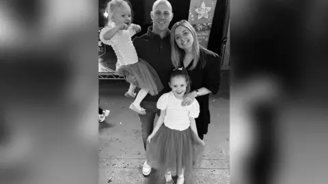 Family handout In this black and white photo, Elsie Dot Stancombe smiles at the camera, wearing a chiffon skirt and pale short sleeved top, with her dad and mum smiling standing behind her and her dad holding Elsie's younger sister Rosie. Rosie is smiling and wearing a matching outfit, holding a toy close to her mouth.