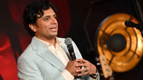 Warner Bros M. Night Shyamalan wearing a white shirt and blue pin-striped blazer