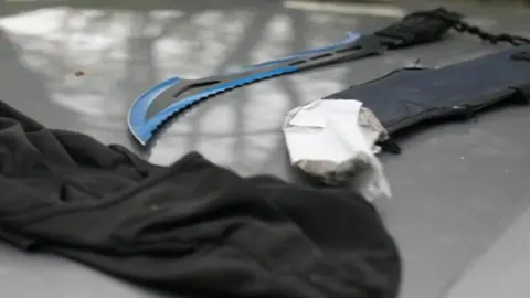 A large curving zombie knife with a bright blue edge lies on the roof of a car that has been stopped by police