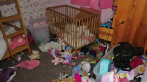 Crown Prosecution Service A very messy child's bedroom, with bunk beds on the left, a cot in the centre and toys and clothes strewn across the floor and a discarded bin bag on the right