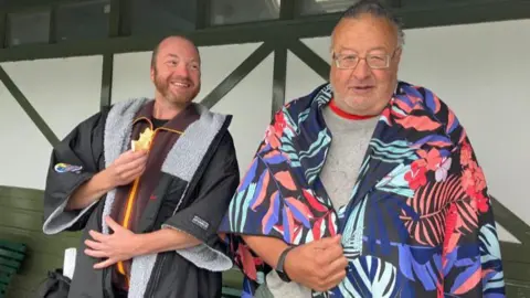 Julia Bryson/BBC William Beebe and Graham Spencer standing in the lido pavilion after their swim, dressed in a towel and a dry robe