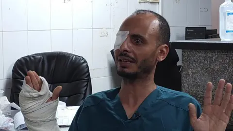 Ashraf El Attar sat down wearing turquoise hospital scrub top and with his hands raised in the air in front of him. His right hand is heavily bandaged and has his fingers and thumb poking out. His right eye is covered in an eye patch