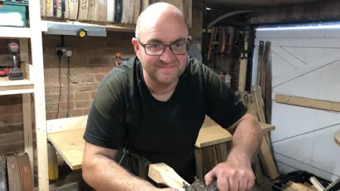 Alan Webber Daniel Garner in his workshop with glasses on and a black t-shirt leaning over wood and smiling into the camera