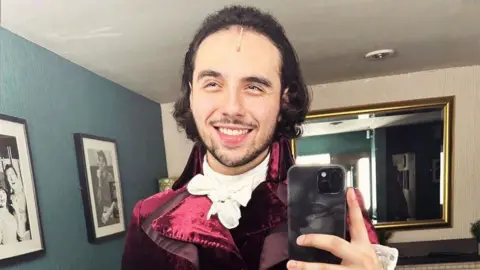 Jonathan Hermosa-Lopez Jonathan Hermosa-Lopez taking a photo of himself in the mirror, while wearing a maroon coloured outfit set in the 1700s