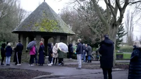 Men and women, some with umbrellas, gather around a stone bandstand with a pointed, tiled roof, on a grey day in Pickering Park, Hull.ull's Pickering Park