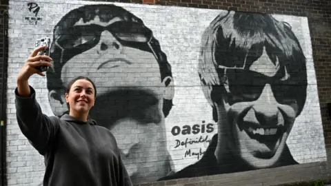 Getty Images Fan Emily McShane, takes a photograph of a new street artwork depicting Oasis' Liam and Noel Gallagher, created by Manchester-based street artist Pic.One.Art. on the side of the Sifters Record store in Burnage, a suburb of Manchester, northern England on August 27, 2024