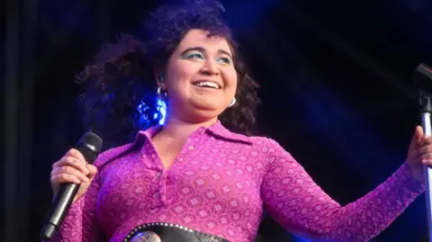 Getty Images Remi Wolf on stage. Remi has curly brown hair and bright blue eye make-up. She wears a patterned pink jumpsuit and smiles into the crowd. 