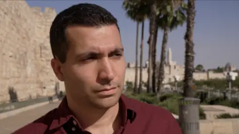 Shai Rosengarten pictured from the shoulders up. He is clean shaven, and wearing a red shirt while looking off camera to the left.