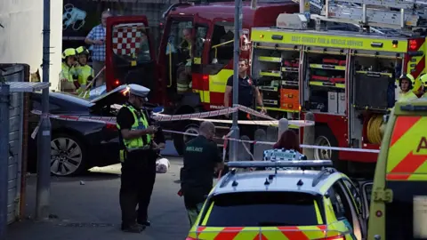 Dan Jessup Emergency service crews surround a black car that has crashed into a white wall. 