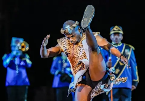 YURI KOCHETKOV/EPA Members of the South African Force Band perform during the opening of the International Military Music Festival 'Spasskaya Tower' on the Red Square in Moscow. In the foreground of the picture is a male dancer energetically kicking his leg up high in the air, wearing a leopard-skin outfit and headdress. In the background are members of the South African Force Band. One is playing a trumpet. They are wearing blue jackets with gold trims.