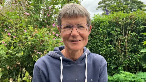 A woman with short grey hair wearing purple circular glasses and a blue drawstring hoodie. She is sitting in front of green bushes and shrubbery. 