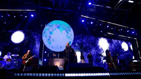 Getty Images Liam Gallagher on stage with musicians and backing singers at the O2 Arena in London. The backdrop is dark blue with a number of large white, fuzzy edged circles and the stage props include a giant globe suspended above them