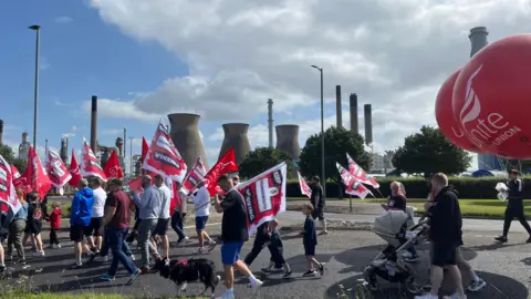 People carrying red and white banners and large red balloons reading Unite the union, marching past the Grangemouth oil refinery - wide range of people including babies in prams and people with dogs