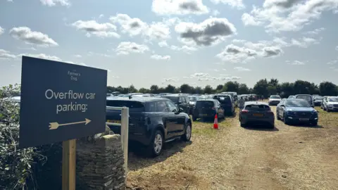 A huge number of cars in a field, with a dark sign for "Overflow car parking"