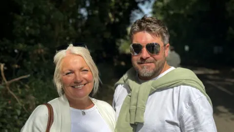 A blonde woman is smiling standing next to her male partner, who is also smiling. 